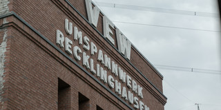Backsteingebäude mit großen weißen Buchstaben an der Fassade, die 'VEW Umspannwerk Recklinghausen' bilden, fotografiert von unten gegen einen bewölkten Himmel.