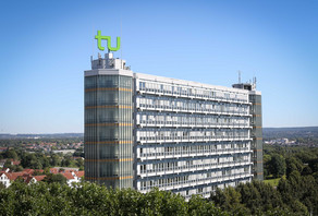 Mathematikgebäude mit Mathetower: Ein hohes Gebäude mit vielen Fenstern und je einem Turm an beiden Seiten. Auf dem linken Turm ein grünes TU-Logo. Im Hintergrund Häuser und Bäume.